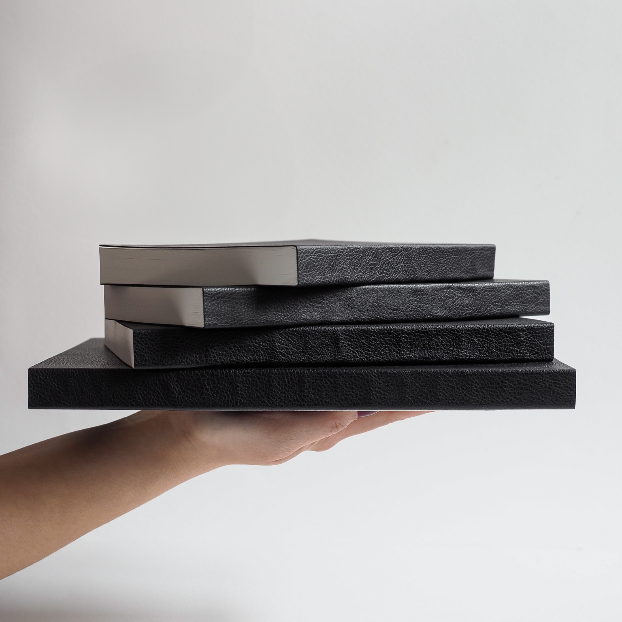 A hand presenting a stack of three sophisticated black journals or notebooks, featuring textured, premium faux leather covers.