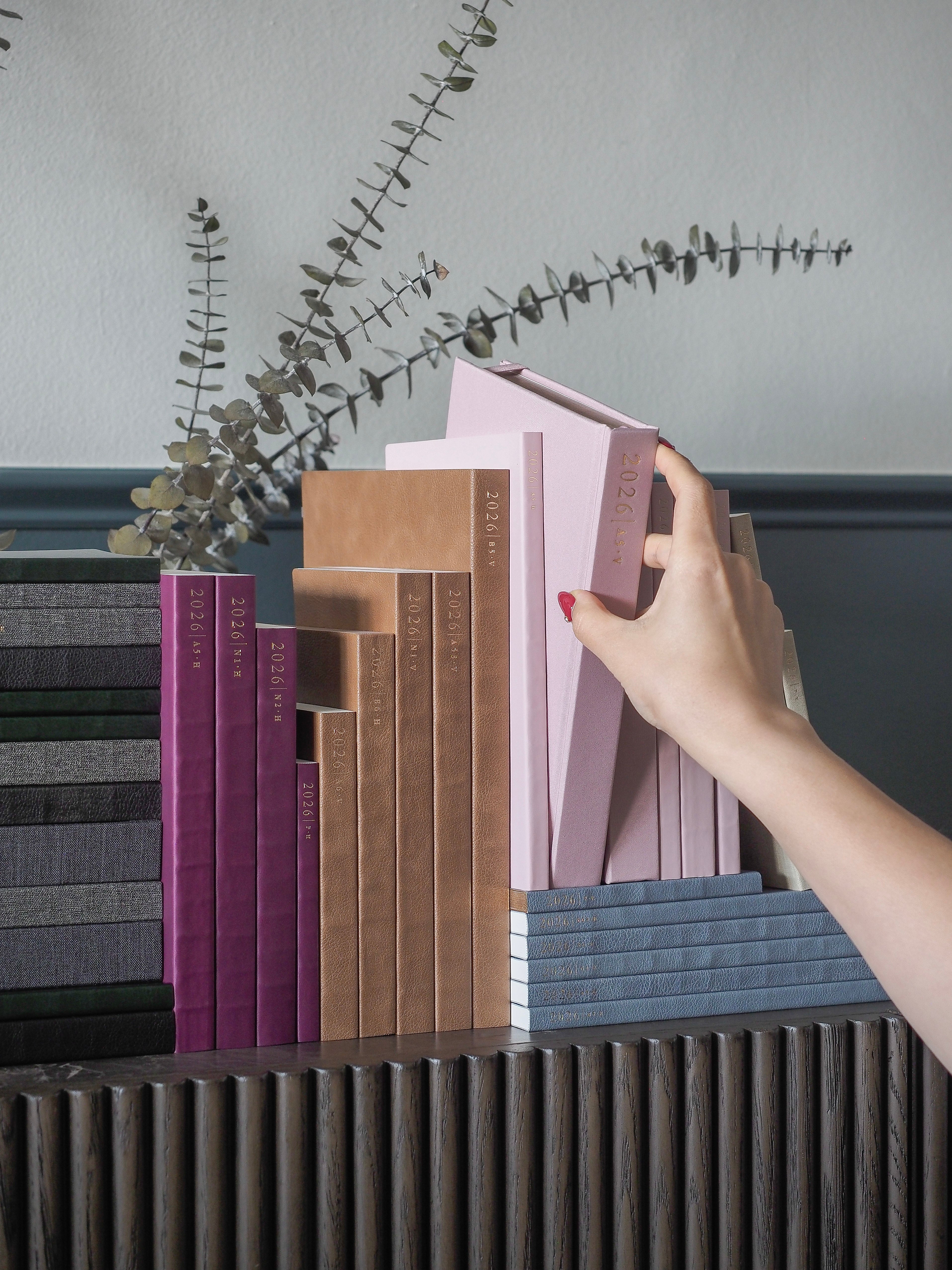 Colorful stack of 2026 planners featuring diverse hues, including deep purple, warm tan, blush pink, and soft blue, presented for organization.