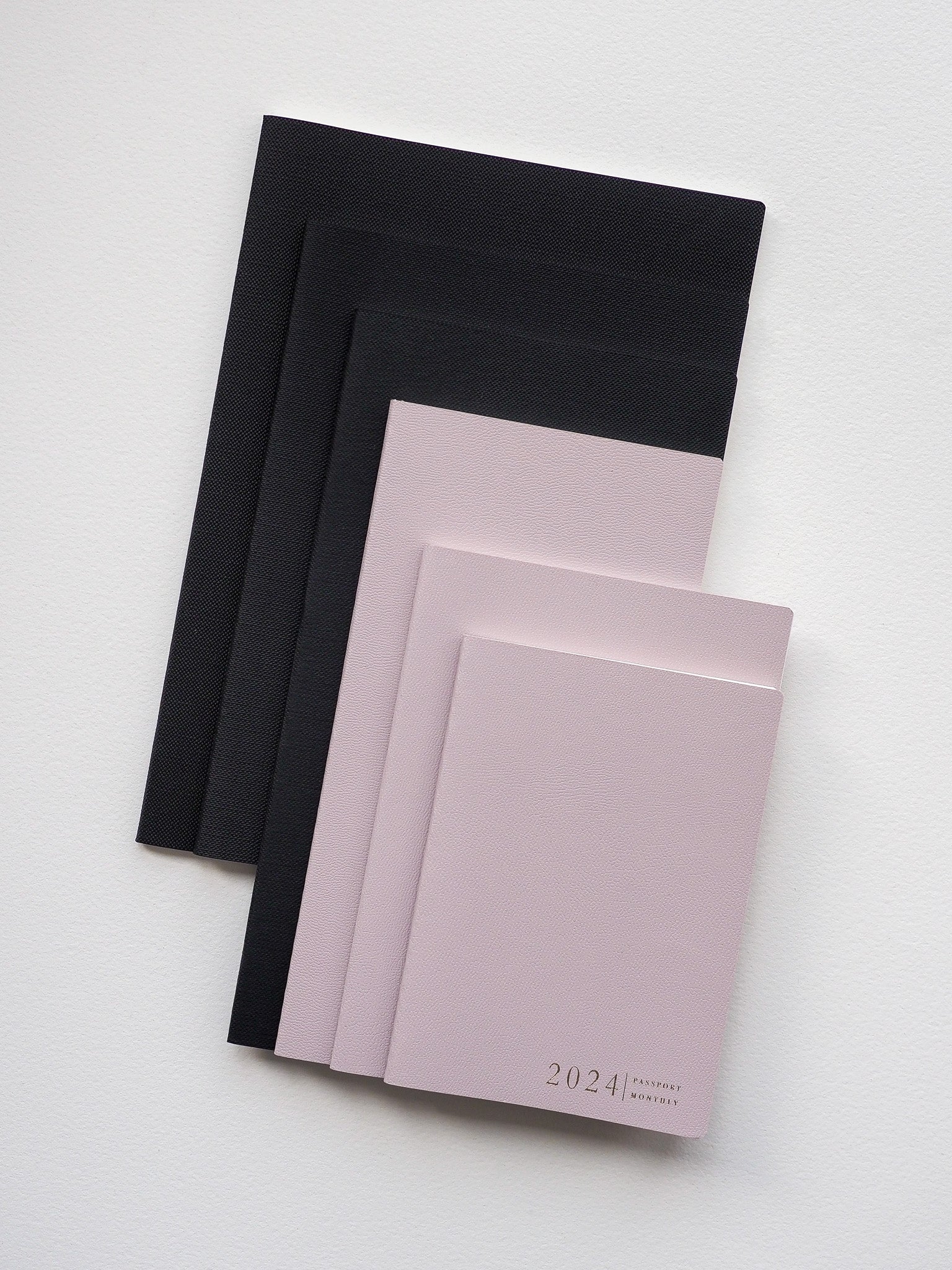 Gradient stack of booklets in black, charcoal, nude, and blush pink tones, with the top planner featuring "2024 Passport Monday" on its cover.