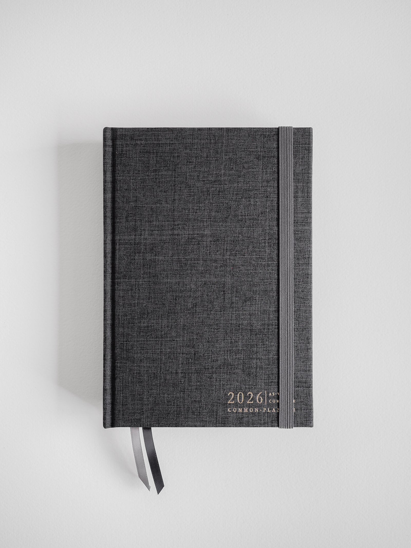 Dark charcoal A5 planner with a linen texture, featuring minimalist gold lettering for "2026 Common Planner" and an attached coordinating bookmark.
