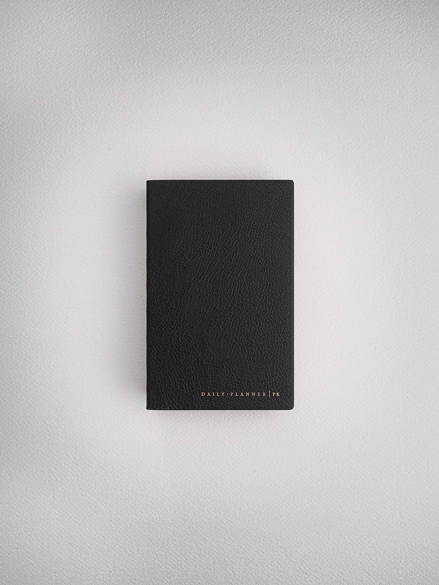 Minimalist black faux-leather daily planner notebook, pocket-sized and debossed with the brand text "DAILY PLANNER | PK," ready for daily organization.