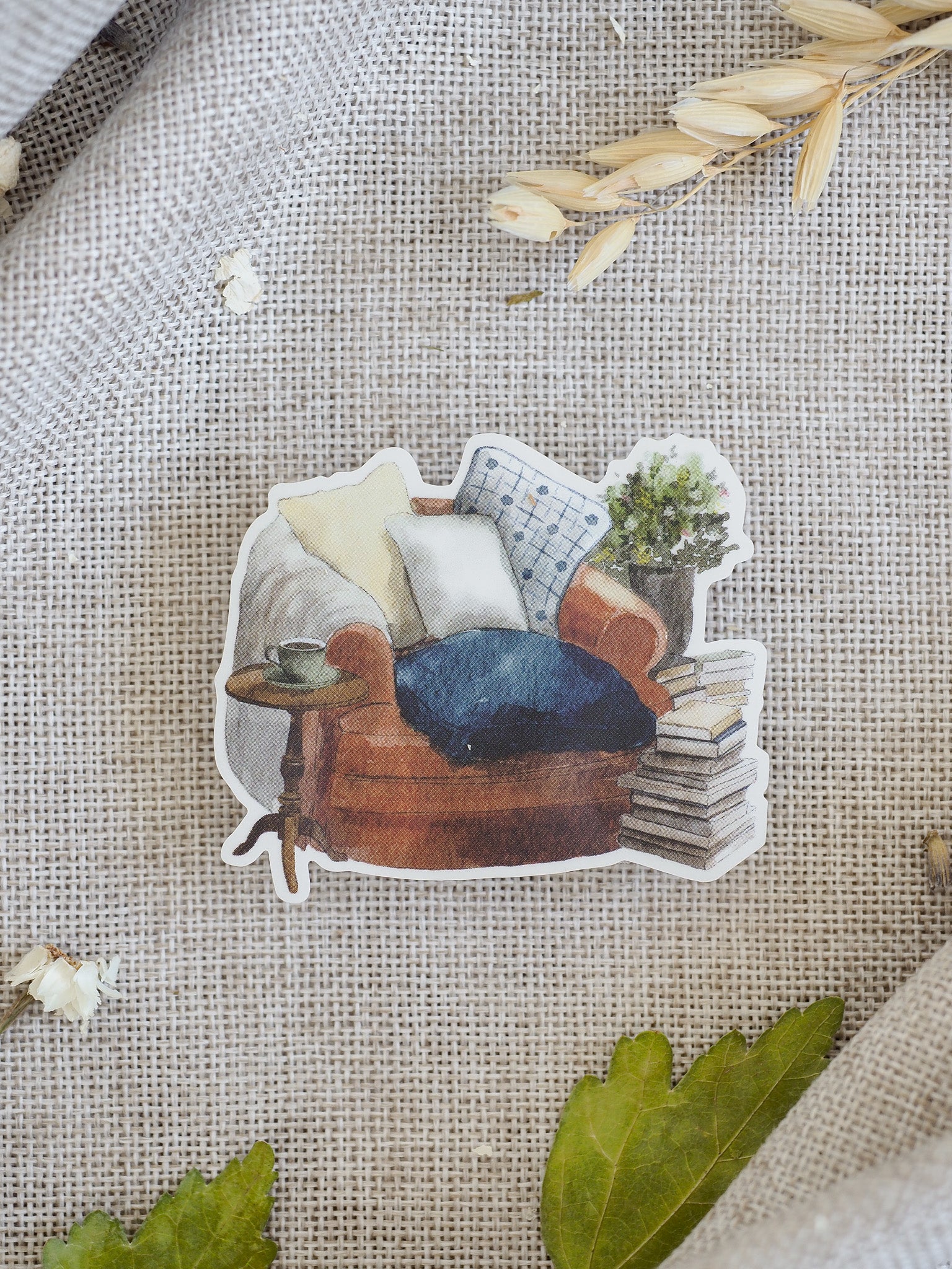 A cozy die-cut sticker depicting a warm brown armchair accented with patterned pillows, stacked books, and a small side table.