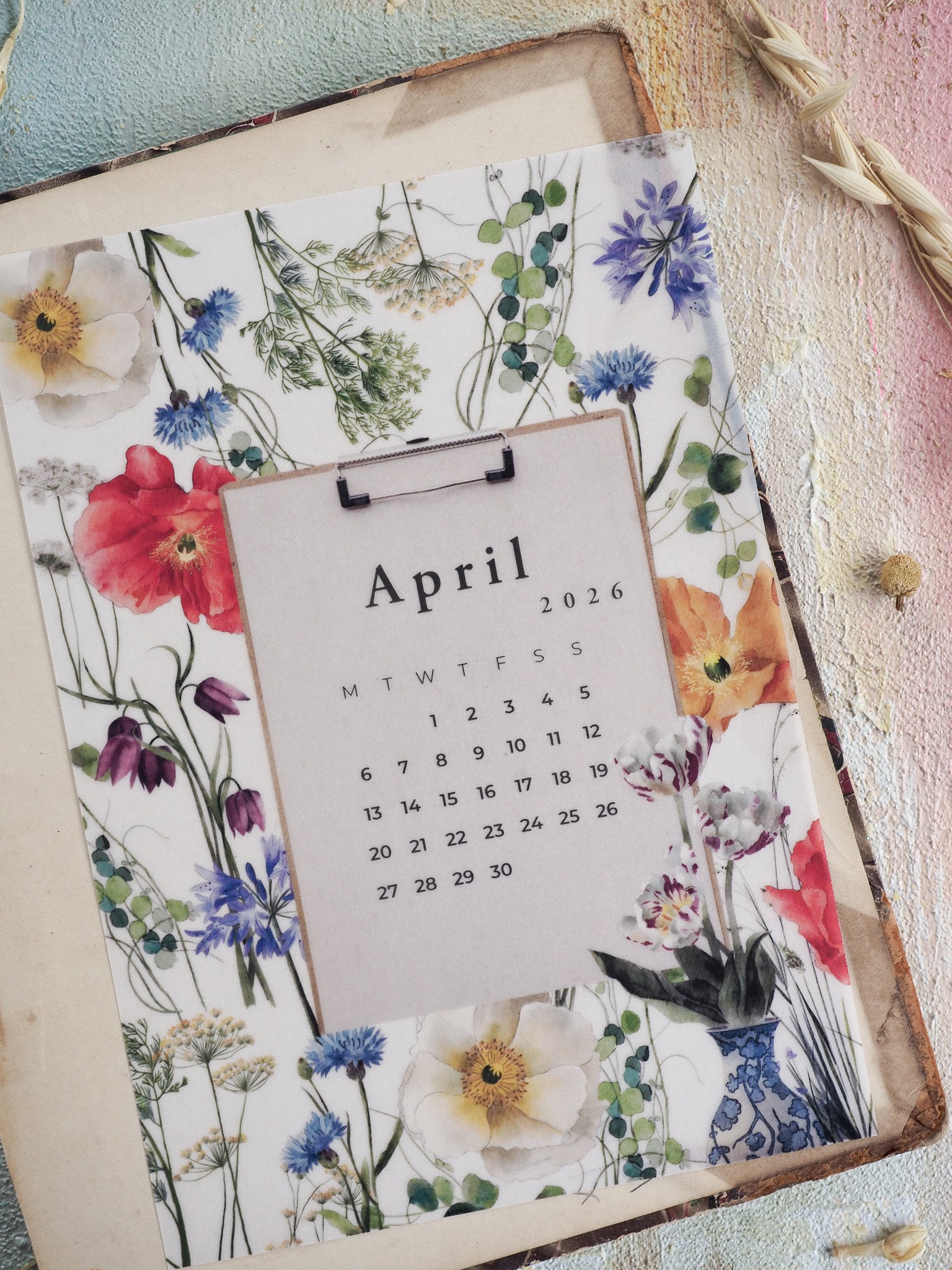 B6 calendar featuring a lush watercolor wreath of spring blooms, including soft yellow, vibrant red, and deep blue flowers, decorated around the April 2026 grid.