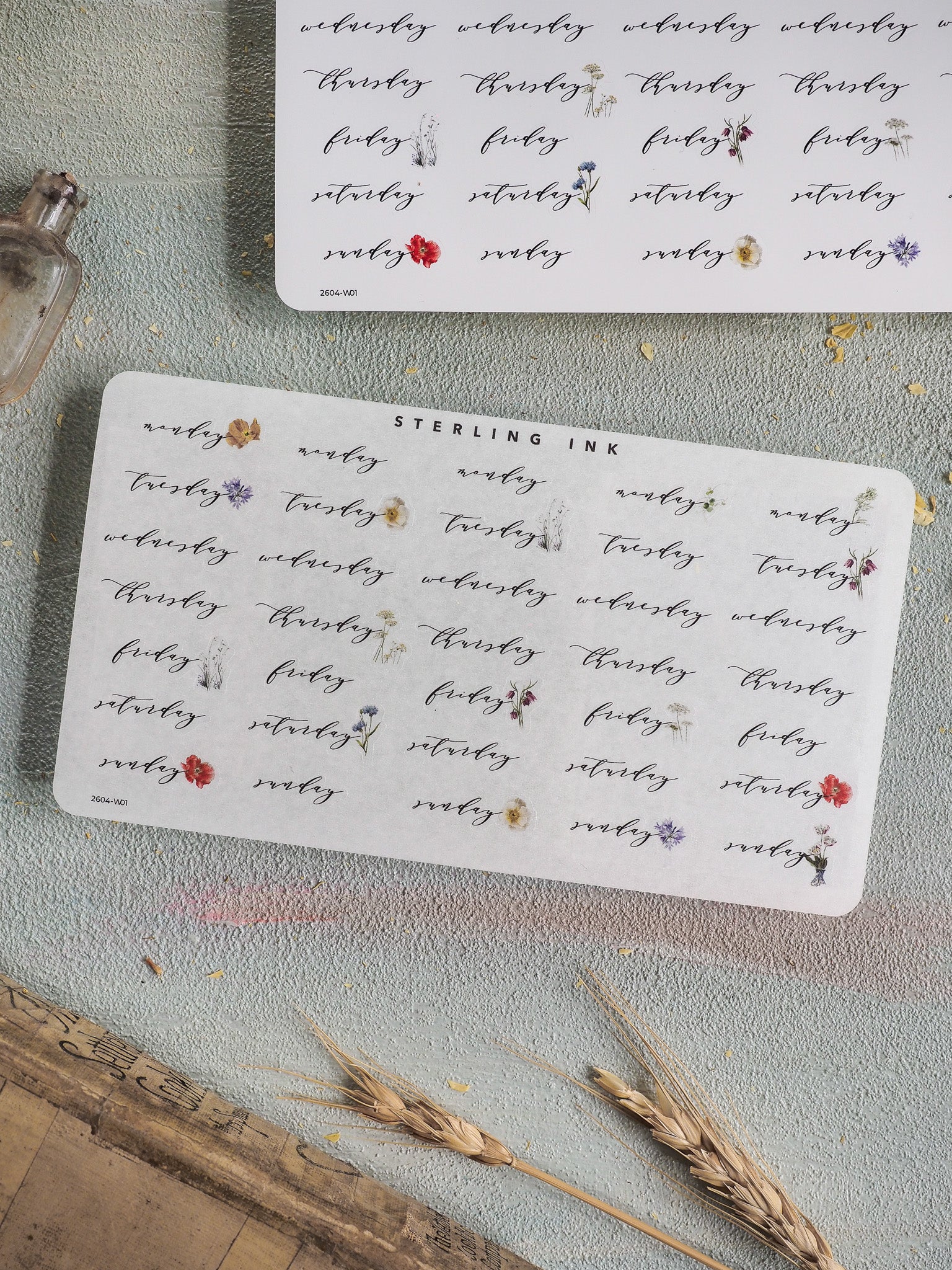 White sticker sheet for weekly planning, featuring delicate printed days of the week interspersed with whimsical red, blue, and purple botanical floral accents.