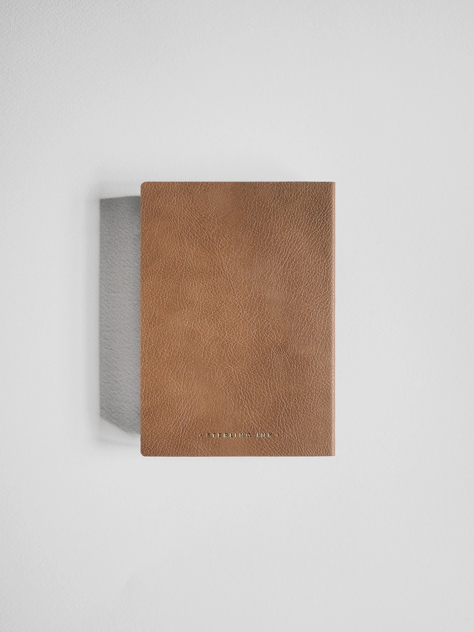 A slim, rectangular brown leather notebook or journal with a textured cover, standing upright against a light gray background.
#Color_Caramel