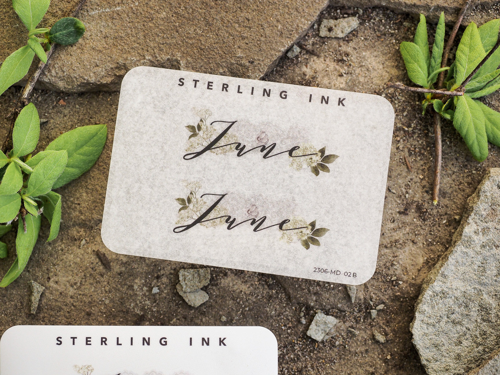 Elegant cream-colored sticker featuring the script word "June" centered with floral accents, framed by the brand name and designed for seasonal journaling.