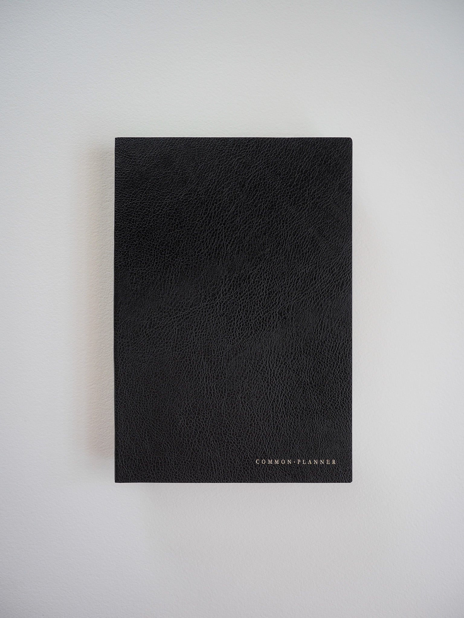 Minimalist matte black A5 planner with a subtly textured cover and the centered, debossed brand name "COMMON: PLANNER."