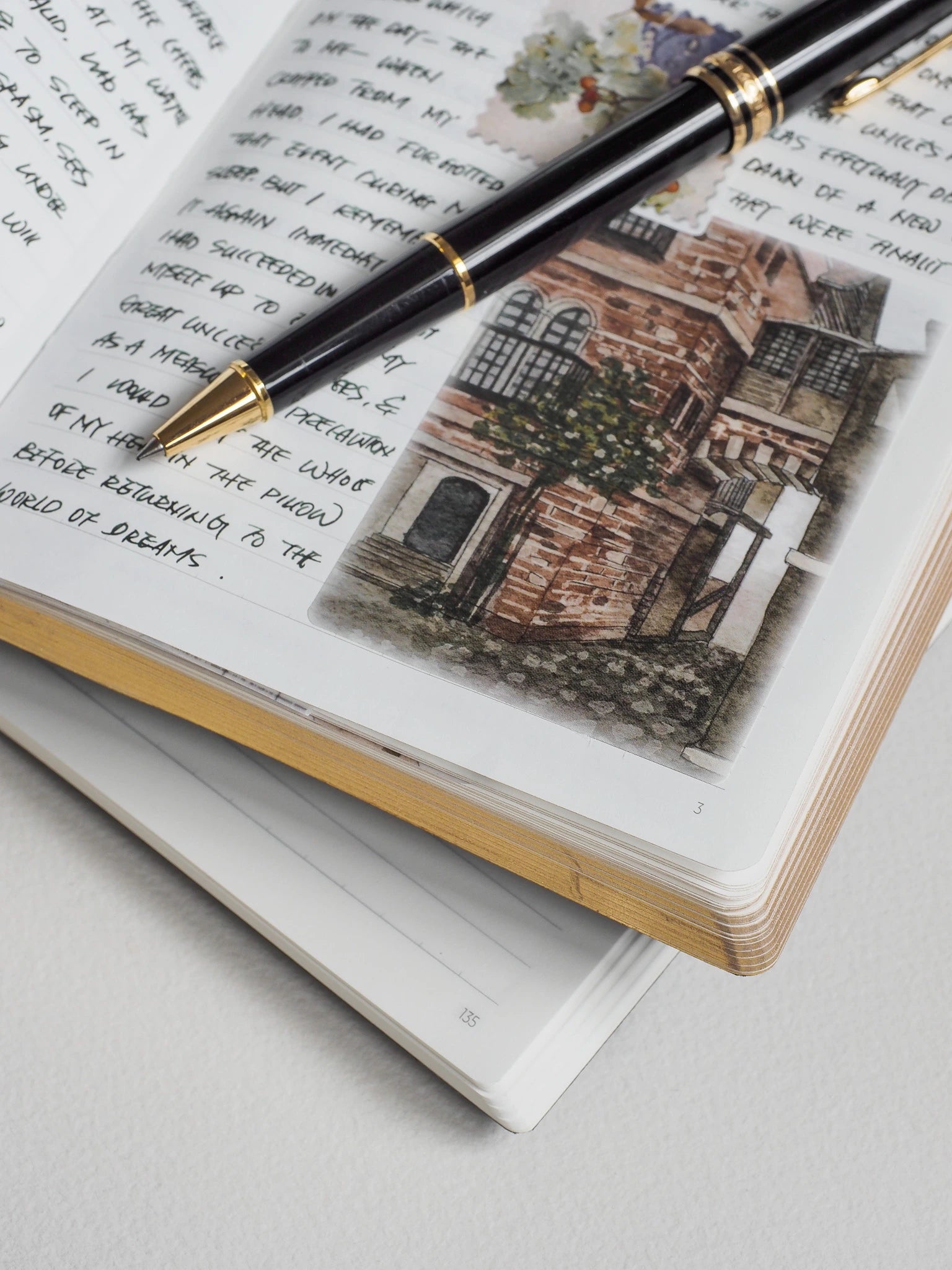 A black fountain pen with gold trim rests diagonally across an open sketchbook. The notebook page features handwritten text and a watercolor illustration of a brick building, with a second notebook page stacked underneath on a light gray surface.