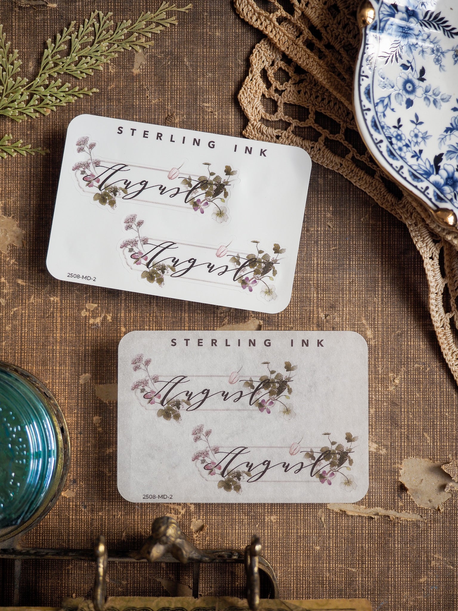 Elegant monthly sticker featuring "August" written in script font, surrounded by delicate watercolor illustrations of mauve and green botanicals.