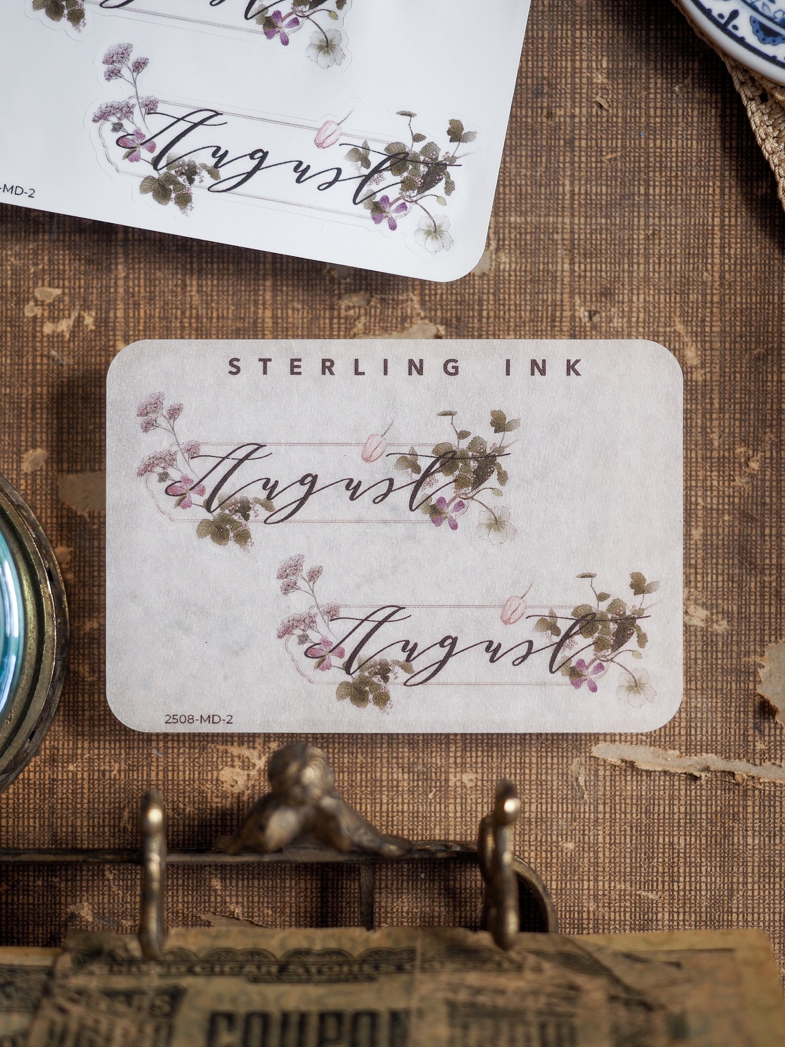 Decorative August month sticker featuring a delicate handwritten script and clustered watercolor floral accents in soft purples and pinks.