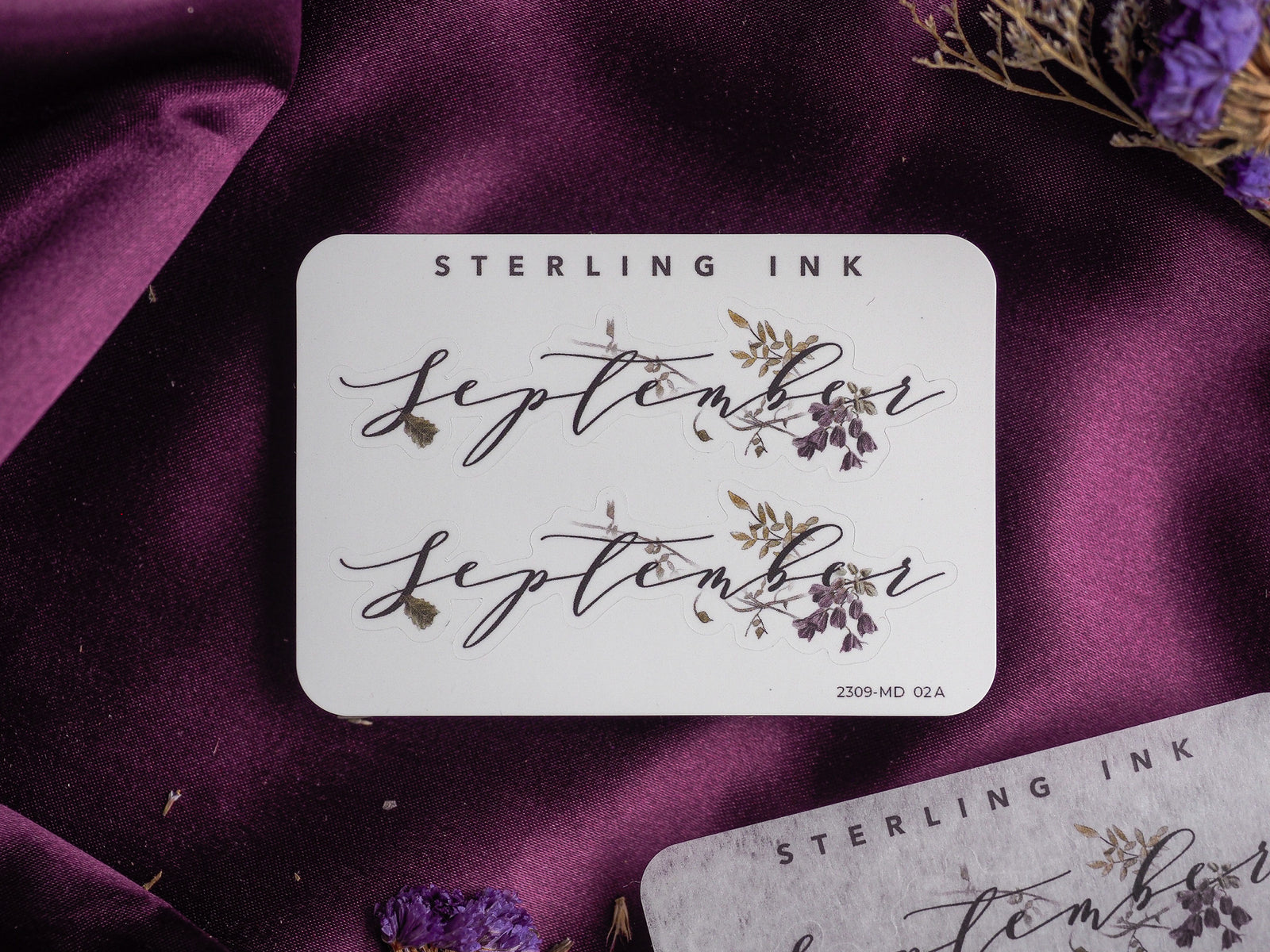 Elegant white month sticker featuring the word "September" in delicate black calligraphy, adorned with sketched natural purple and green floral accents.