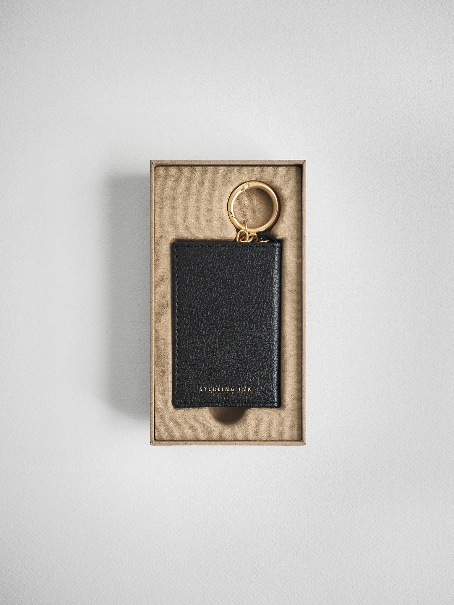 A black textured leather keychain with a gold-tone key ring, presented inside a beige cardboard gift box on a light gray background.
#Color_Onyx