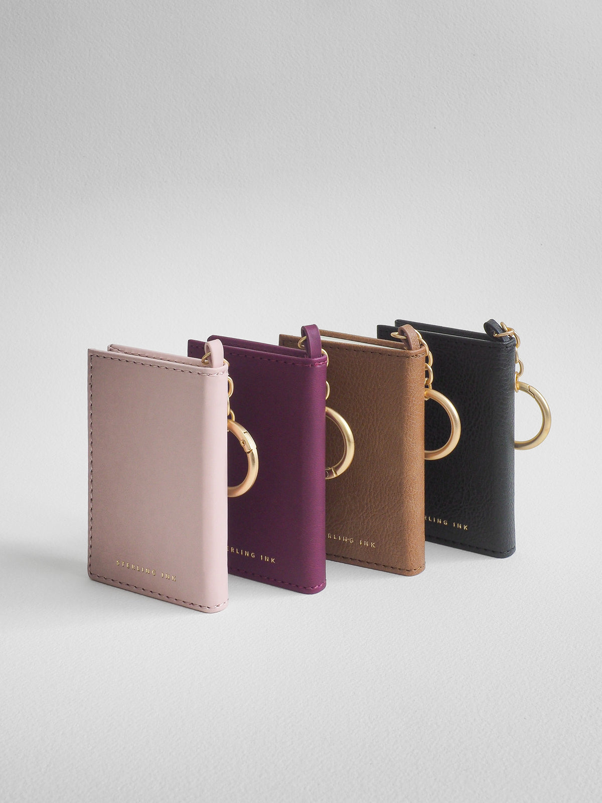 Four slim leather card holder/key case keychains in dusty pink, plum purple, tan brown, and black, arranged in a row on a light gray background. Each case features a gold-tone metal key ring and a small gold logo at the bottom.