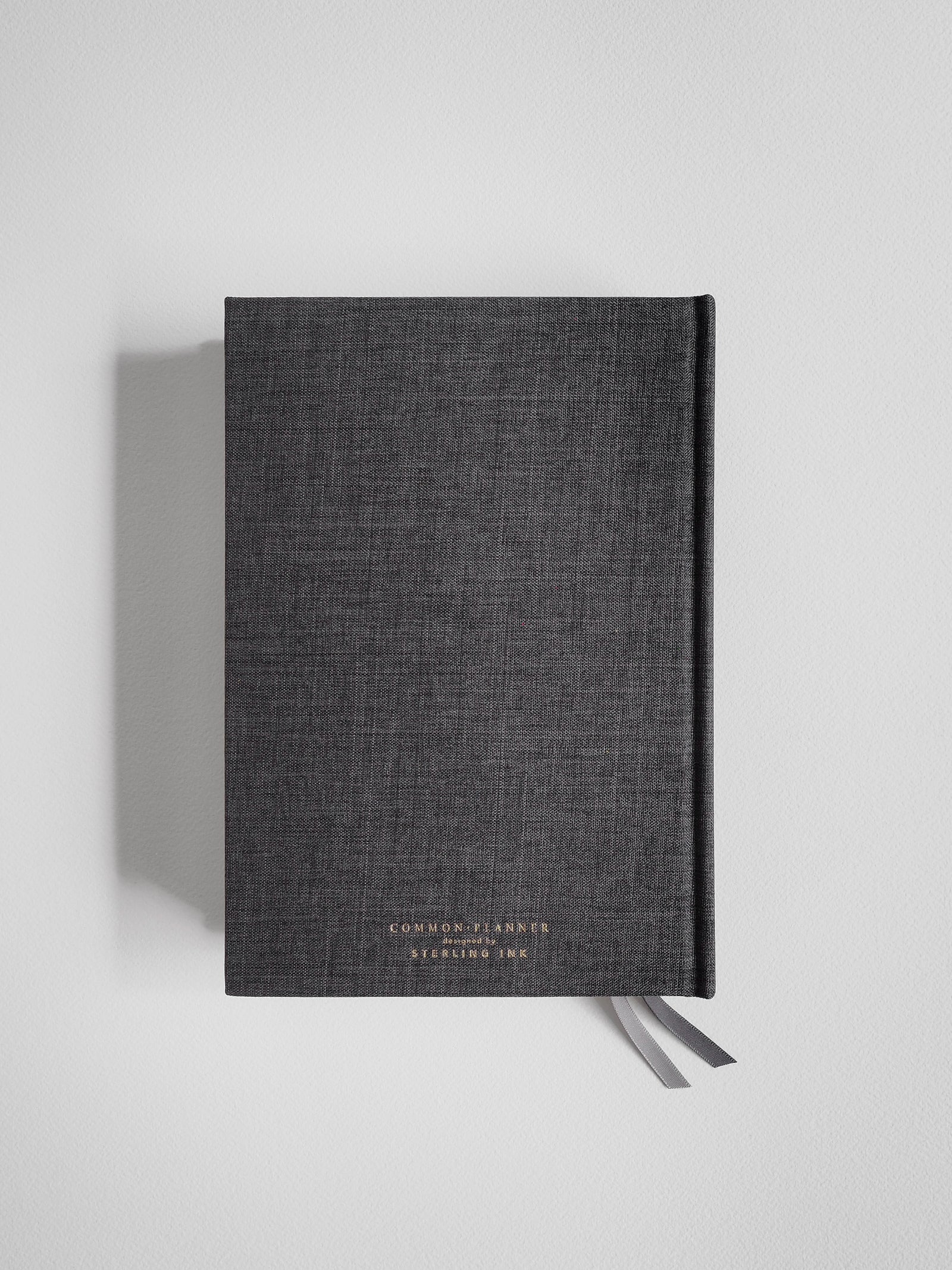 A dark gray linen-covered notebook with a textured cover, featuring small gold foil branding text near the bottom and a matching gray ribbon bookmark, shown against a light gray background.
#Color_Charcoal