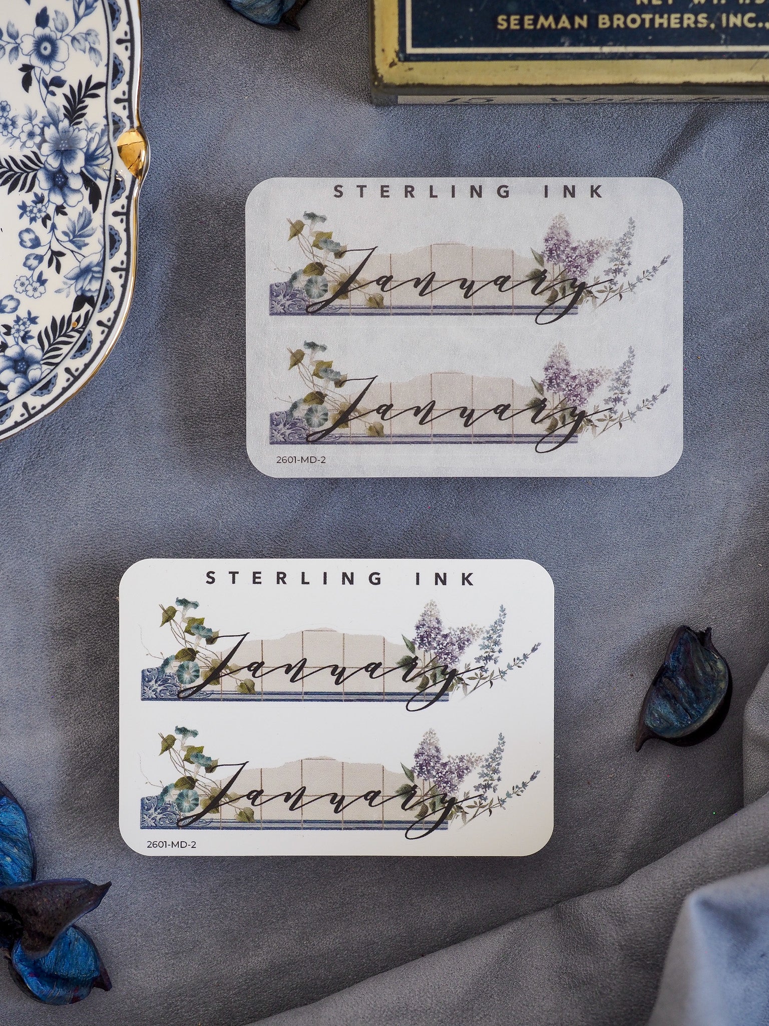 Elegant "January" month sticker featuring scripted text over a delicate watercolor illustration of blue foliage and purple wildflowers.