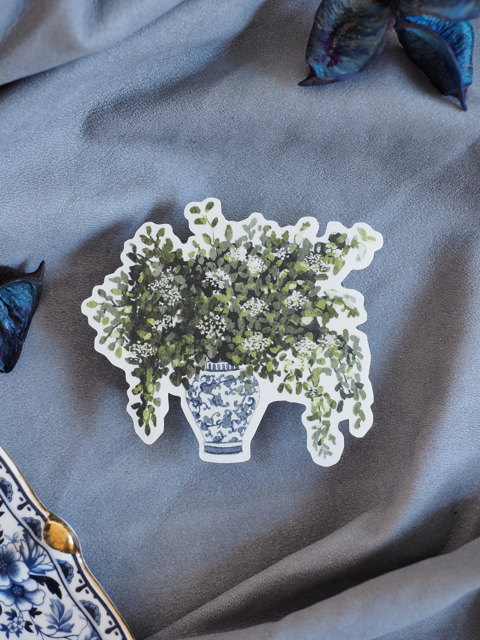 Depiction of a blue and white patterned flower pot overflowing with abundant, verdant green and white floral arrangements.
