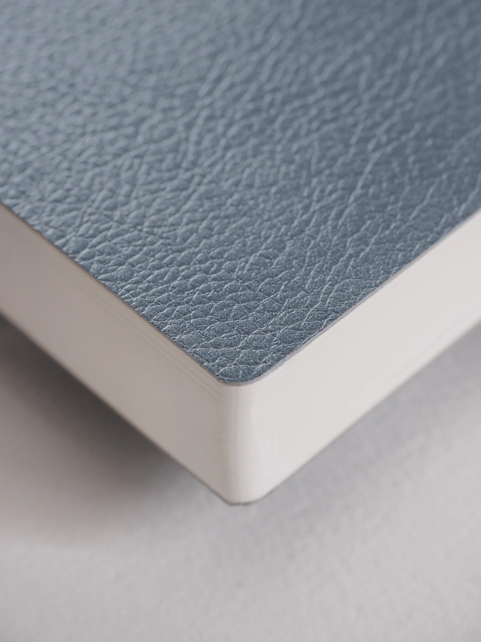 Close-up of a notebook or book corner showing a textured dusty blue cover and white page edges on a light gray surface.
#Color_Mist