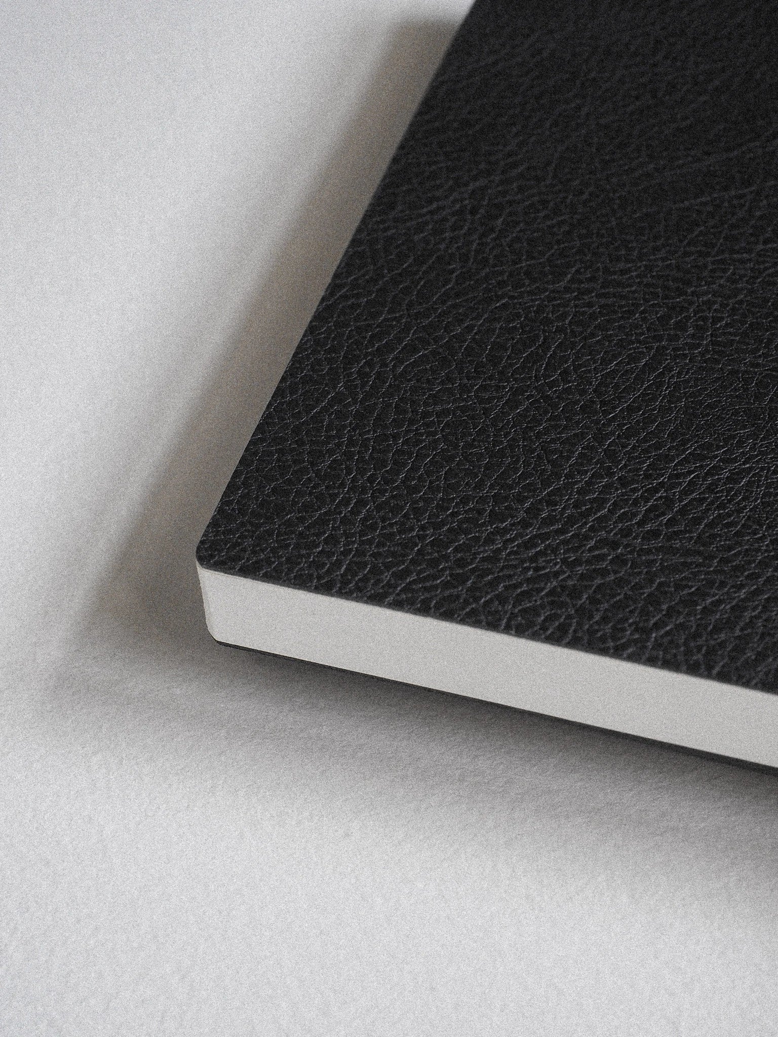 A close-up of a black textured notebook or journal corner with white page edges, resting on a light gray surface.
#Color_Onyx