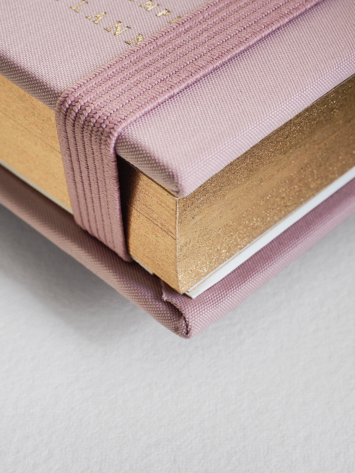 A close-up of a hardcover notebook or journal with a dusty pink fabric cover, a gold glitter-textured page block, and a matching pink elastic closure band.
#Color_Seashell