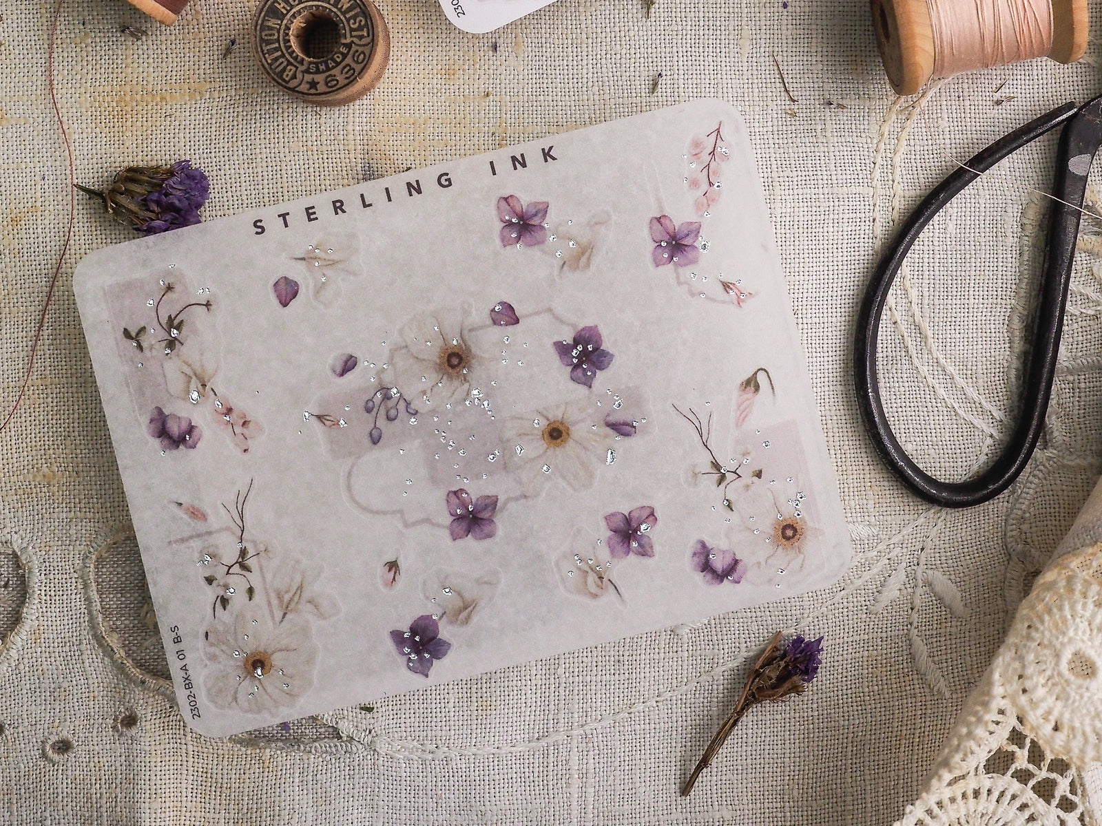Delicate sticker sheet displaying a subtle pattern of watercolor purple and white flowers accented by scattered silver foil details, branded "STERLING INK."