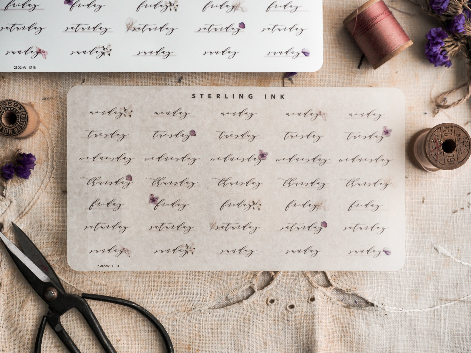 Creamy-white planner stickers featuring repeated, hand-lettered days of the week (Monday through Sunday) and small delicate purple floral accents.