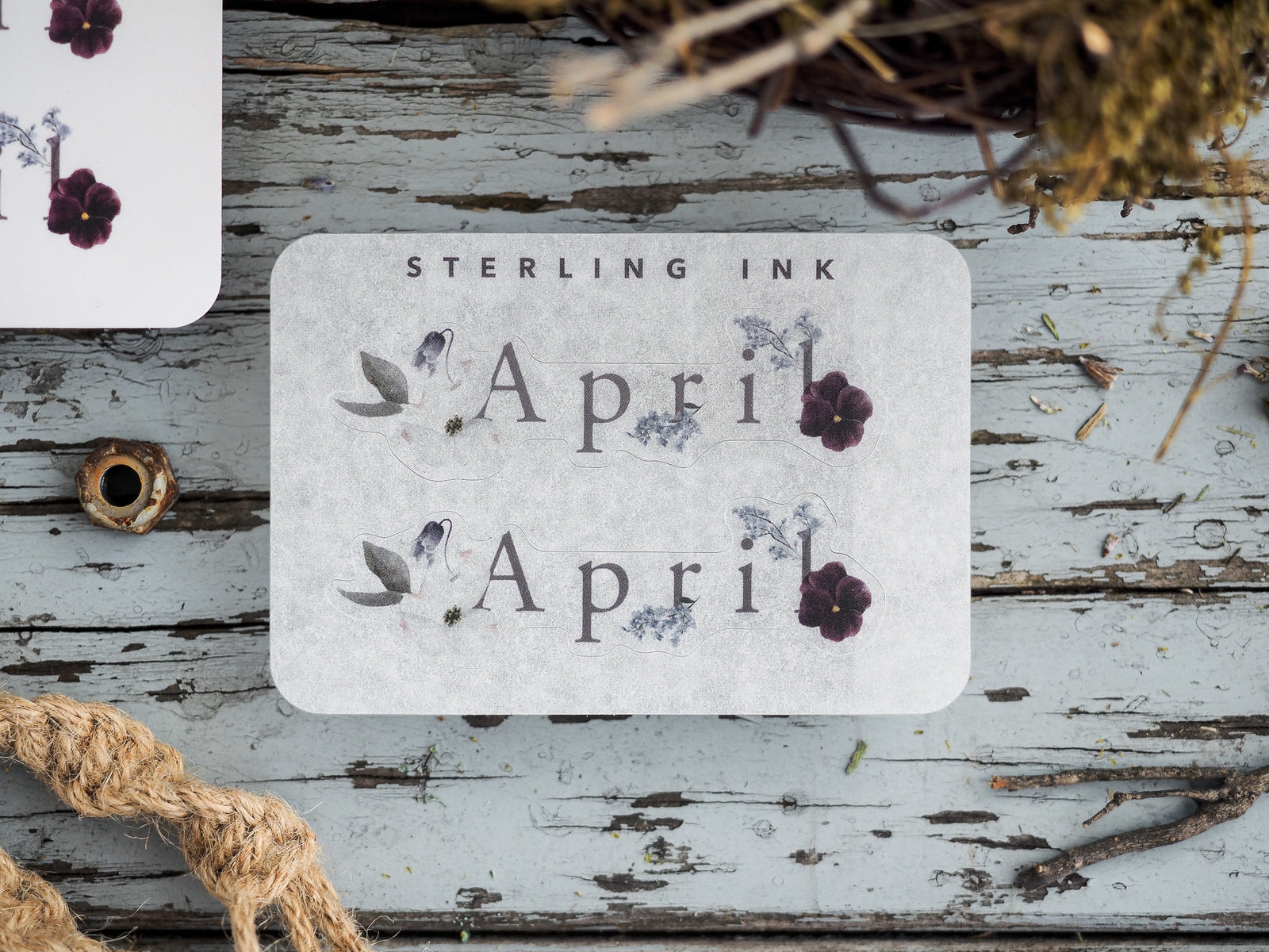 Elegant botanical sticker for April 2023 featuring the word "April" in serif text and delicate decorative accents of purple flowers and sepia-toned leaves.