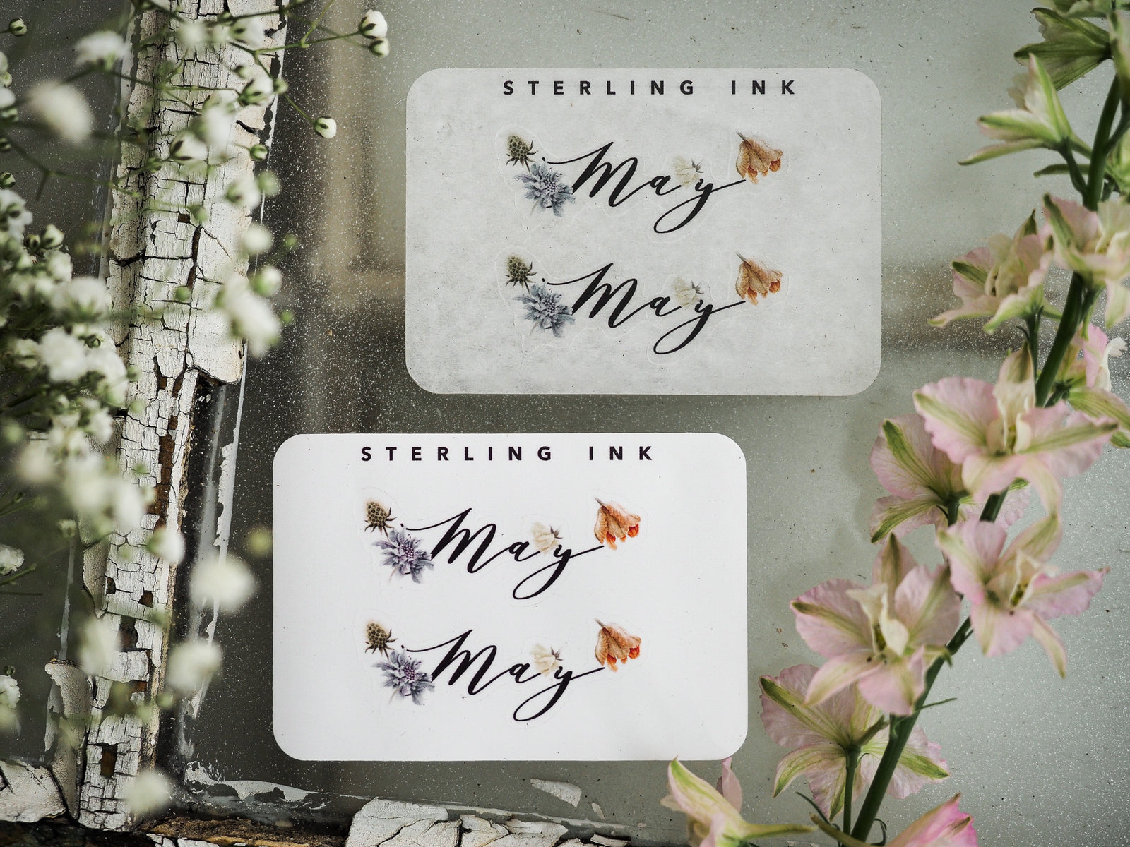 Two white monthly stickers for May, featuring the script word "May" decorated with delicate floral clusters of lavender and peach accents.