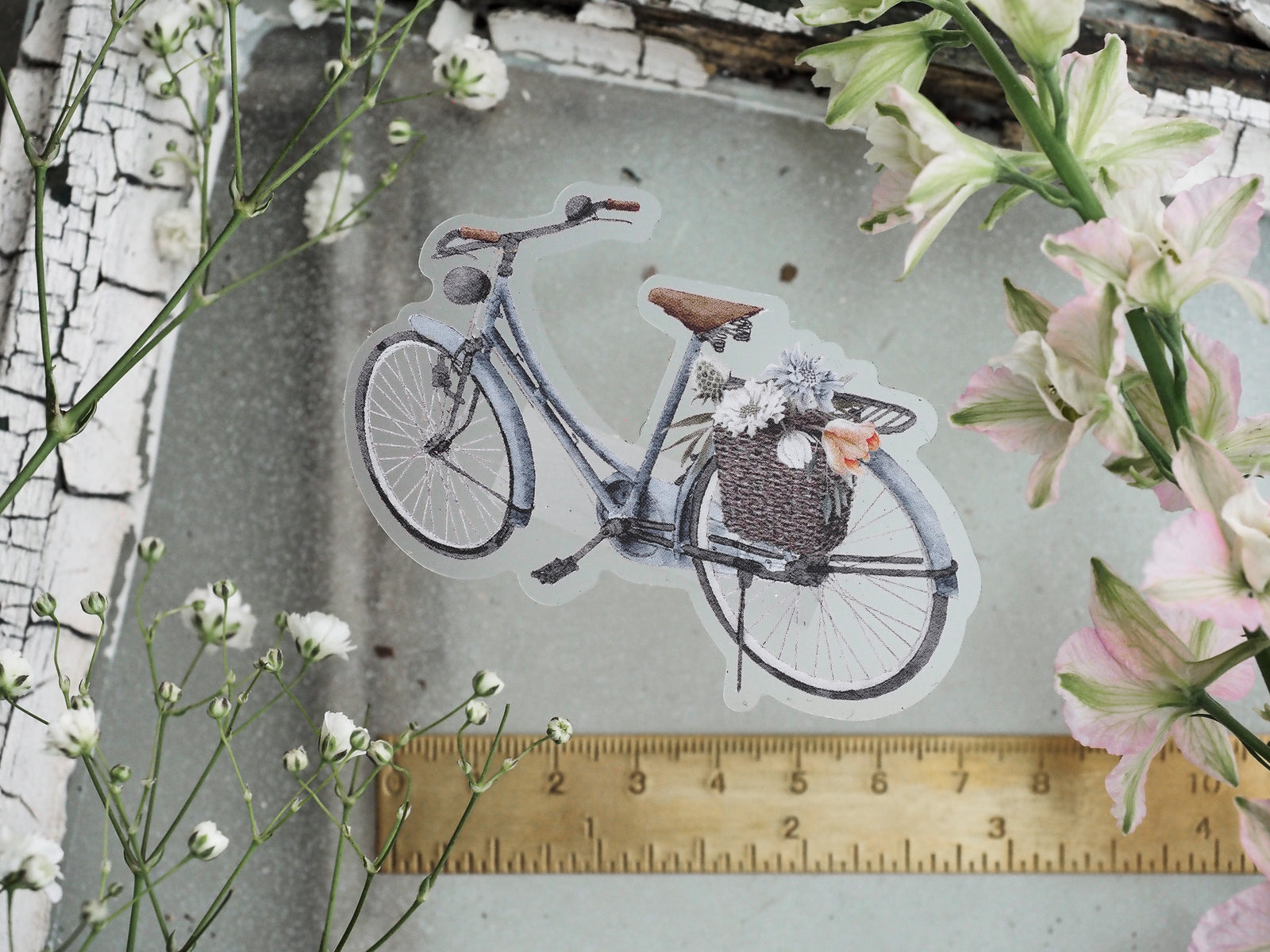 Vintage die-cut sticker featuring an artistic, monochromatic bicycle carrying a rustic basket filled with detailed spring blooms.