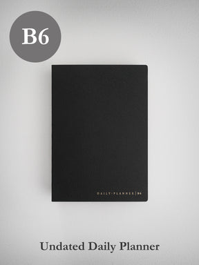 A matte black B6 daily planner journal featuring a structured, textured cover and subtle gold-toned branding near the bottom.