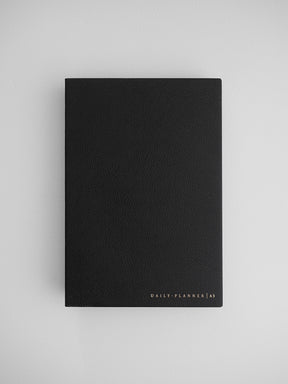 Elegant black A5 daily planner journal with a matte, textured cover, featuring subtle gold metallic lettering that reads "DAILY PLANNER | A5".