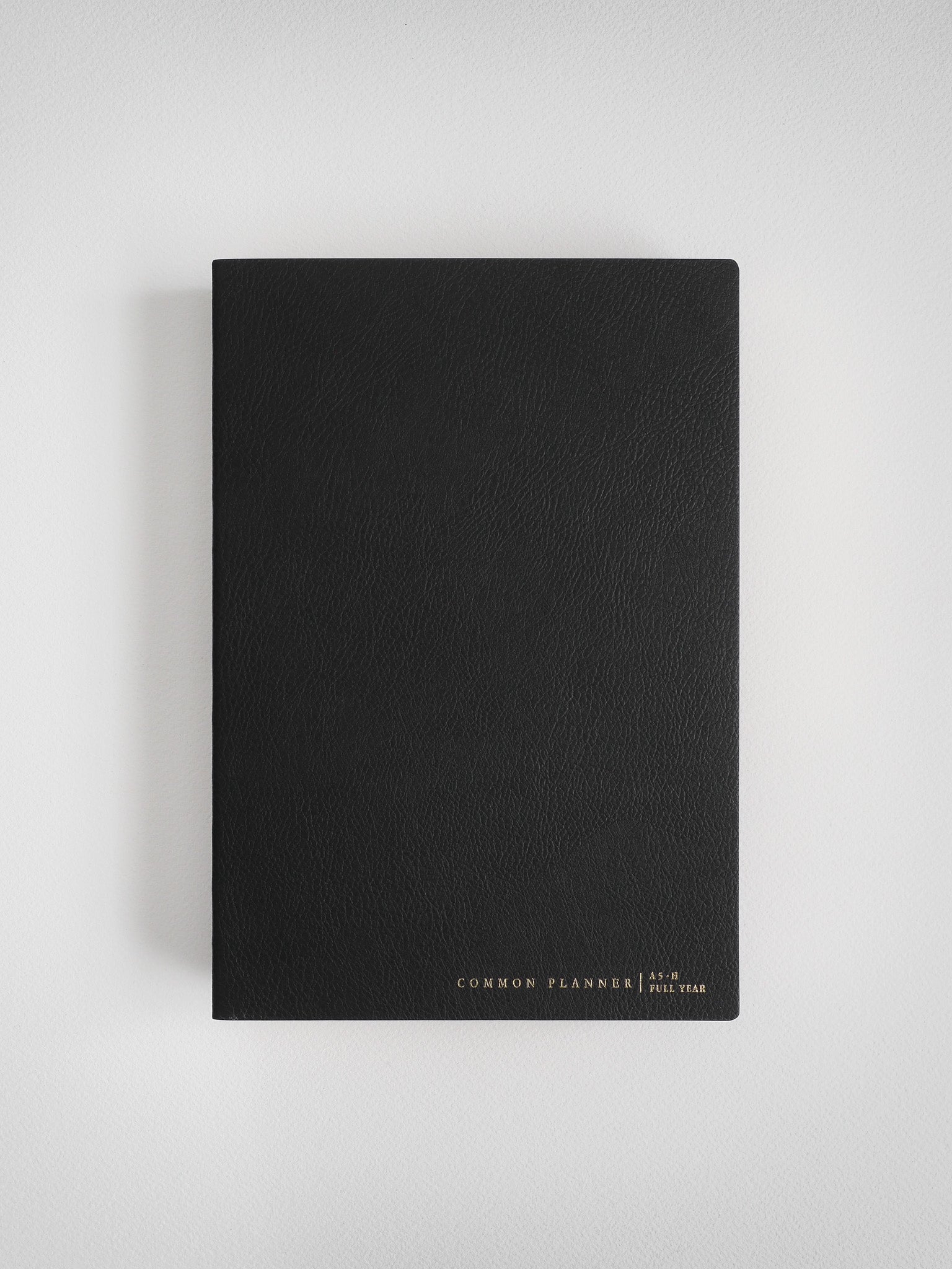 A minimalist black A5 common planner notebook with a matte cover, featuring subtle gold-foil lettering identifying it as a full-year organizational planner.