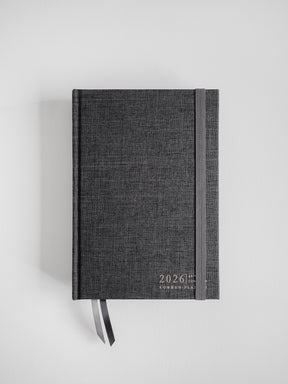 Dark charcoal A5 planner with a linen texture, featuring minimalist gold lettering for "2026 Common Planner" and an attached coordinating bookmark.