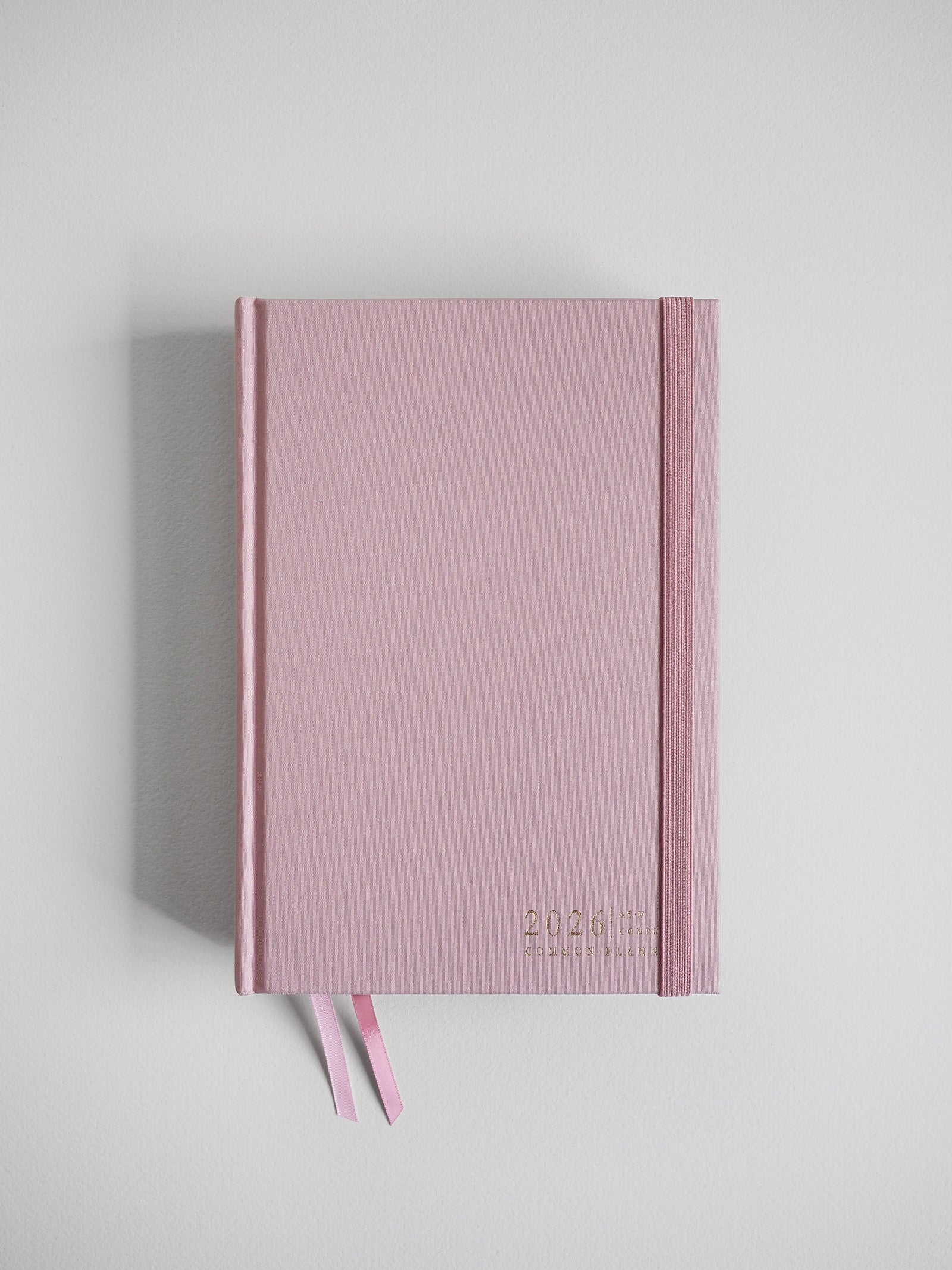 A blush pink, textured A5 planner for 2026, featuring the date and brand title, complete with two coordinating ribbon bookmarks.