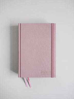 A blush pink, textured A5 planner for 2026, featuring the date and brand title, complete with two coordinating ribbon bookmarks.