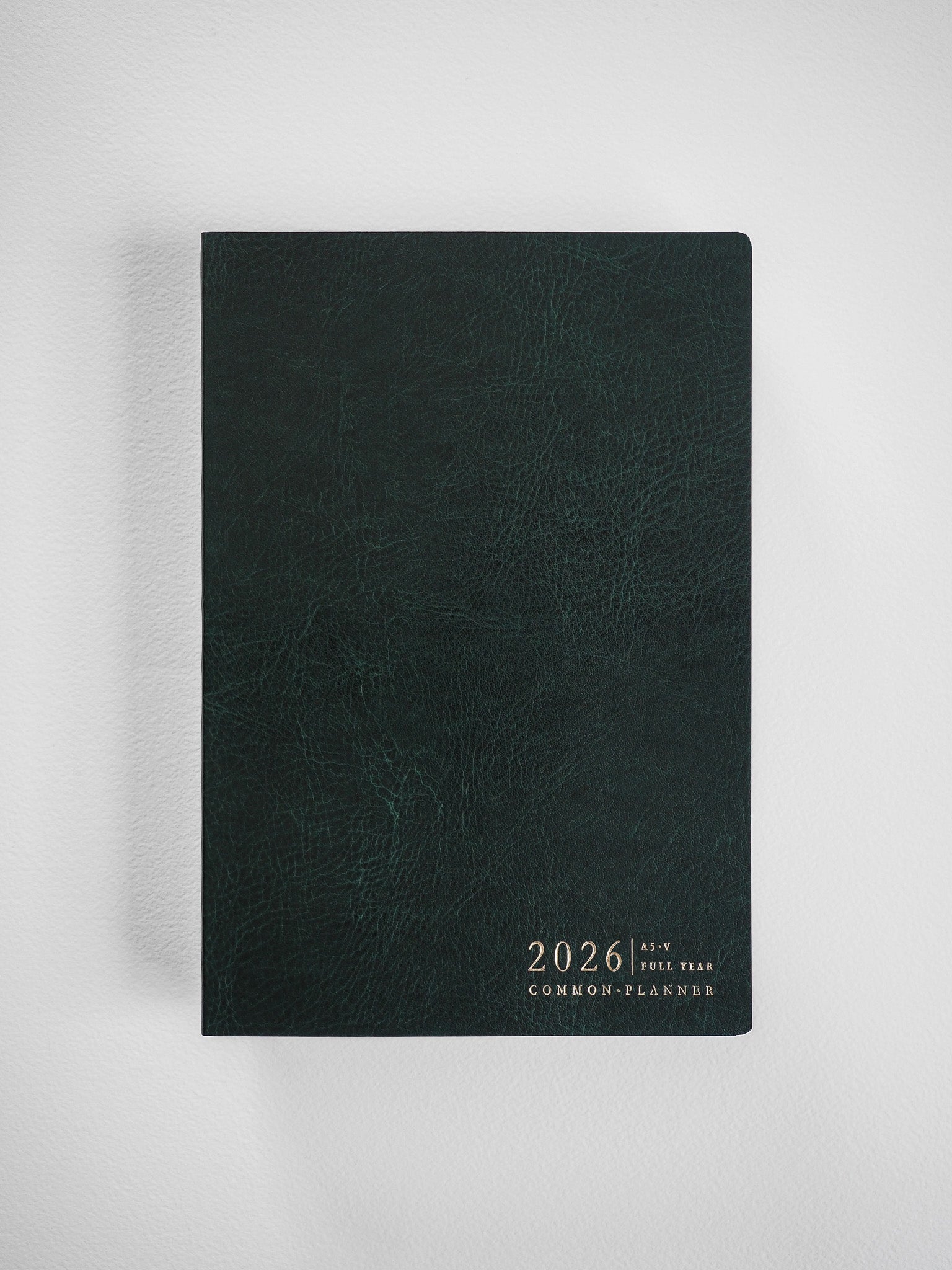 Deep forest green A5 planner with a textured, synthetic leather cover displaying the embossed title "2026 Full Year Common Planner."