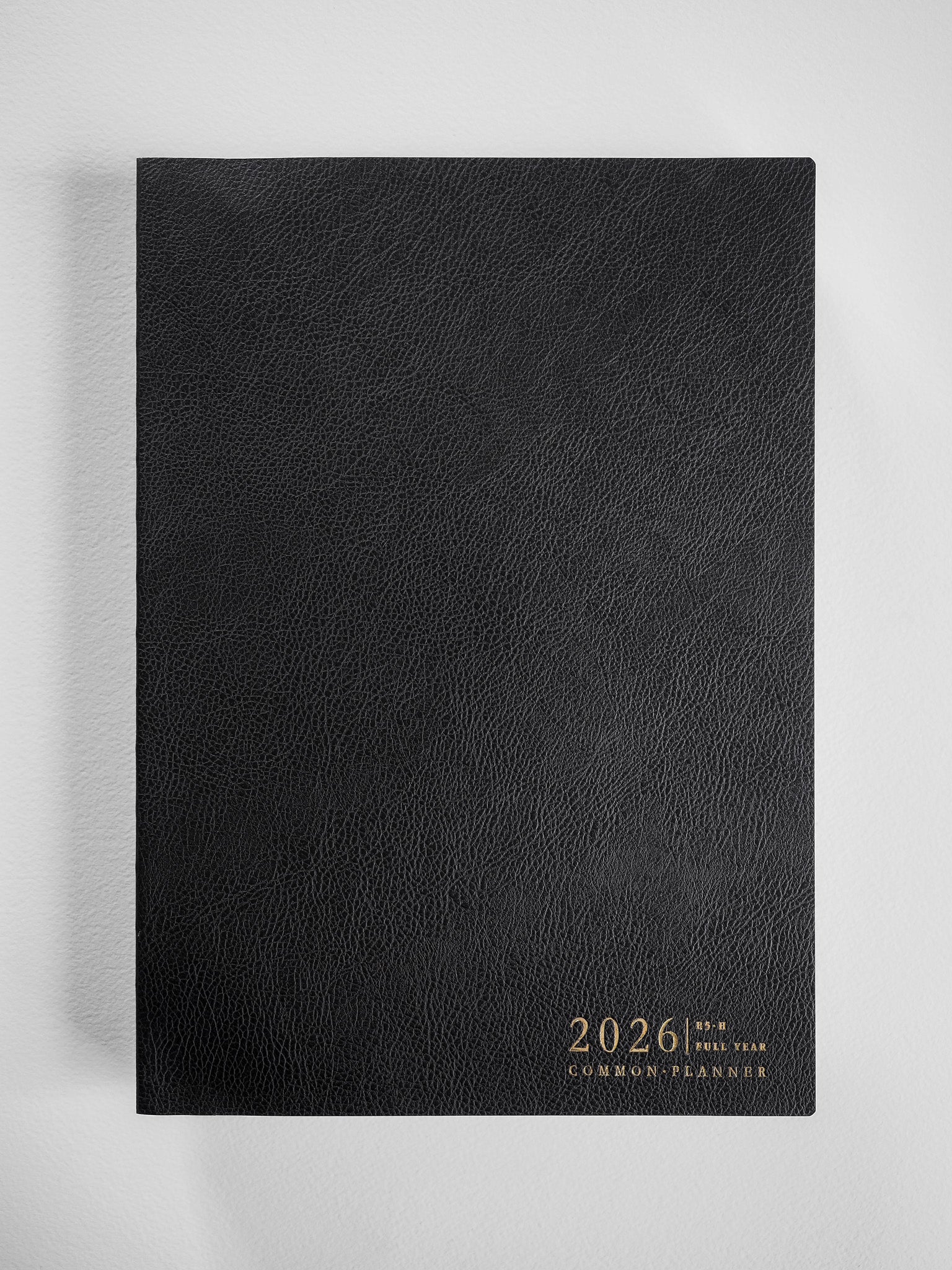 A black, textured B5 planner featuring subtle gold-toned embossed lettering that reads "2026 | FULL YEAR COMMON PLANNER."