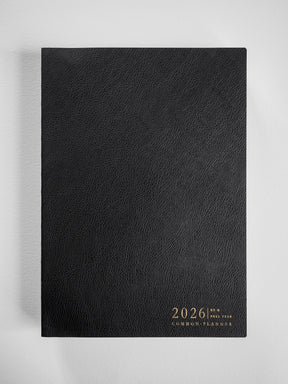 A black, textured B5 planner featuring subtle gold-toned embossed lettering that reads "2026 | FULL YEAR COMMON PLANNER."