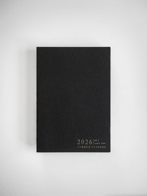 Sleek matte black B6 vertical planner cover adorned with sophisticated gold metallic foil lettering announcing the 2026 Common Planner.