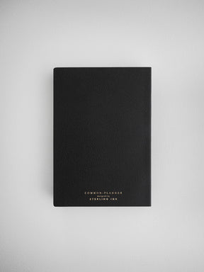 Sleek black planner notebook with a faux leather texture, featuring embossed gold lettering that reads "COMMON PLANNER designed by STERLING INK."