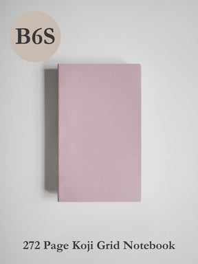 A minimalist pink notebook with a grid layout, shown closed on a light gray background. Text overlays indicate 'B6S' in a beige circle at the top left and '272 Page Koji Grid Notebook' at the bottom.
#Color_Fairyfloss