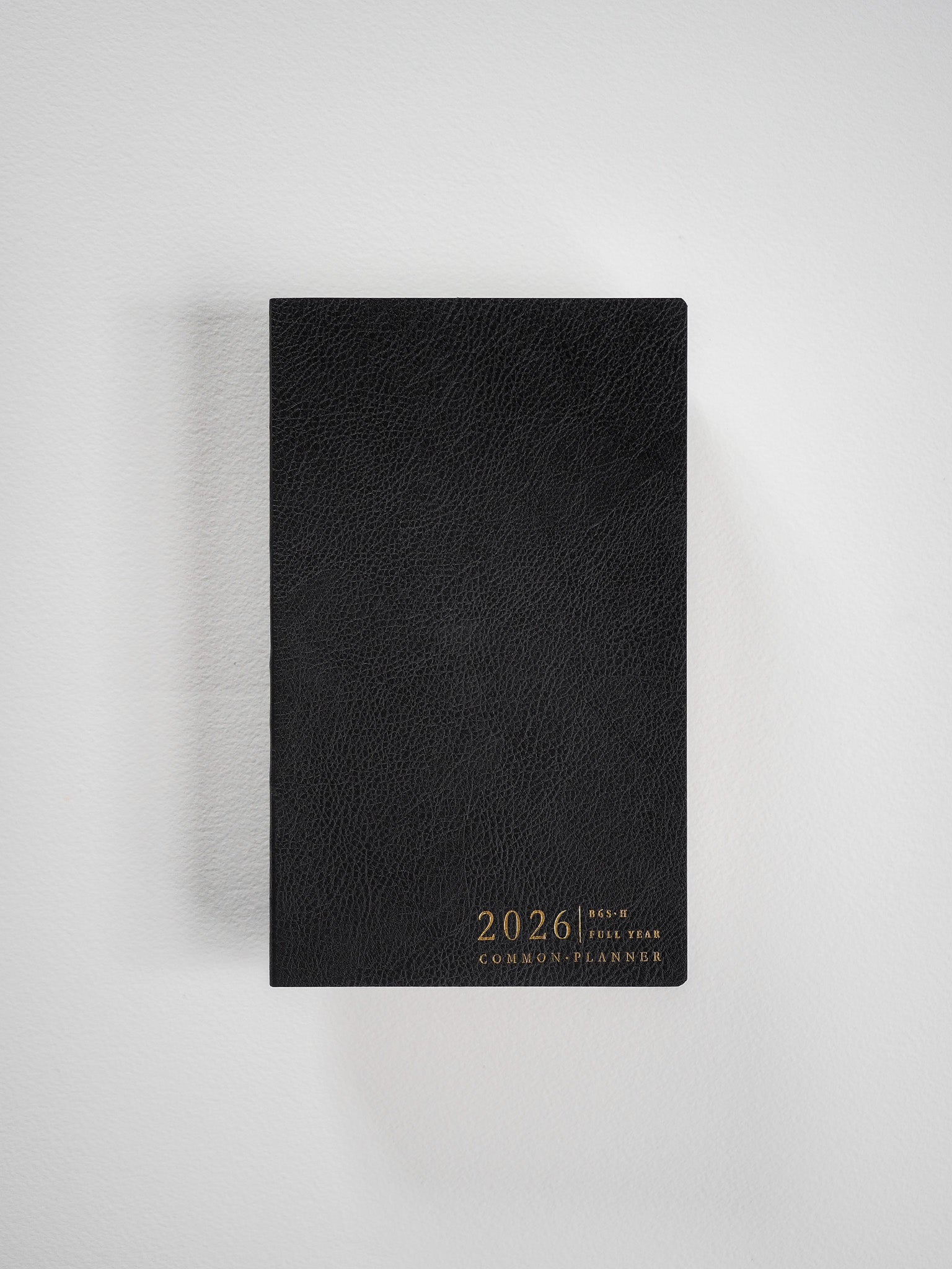 A sleek, black B6 slim planner featuring a textured cover and elegant metallic gold lettering that displays "2026 Full Year Common Planner."