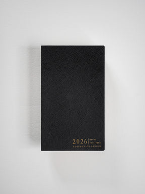 A sleek, black B6 slim planner featuring a textured cover and elegant metallic gold lettering that displays "2026 Full Year Common Planner."
