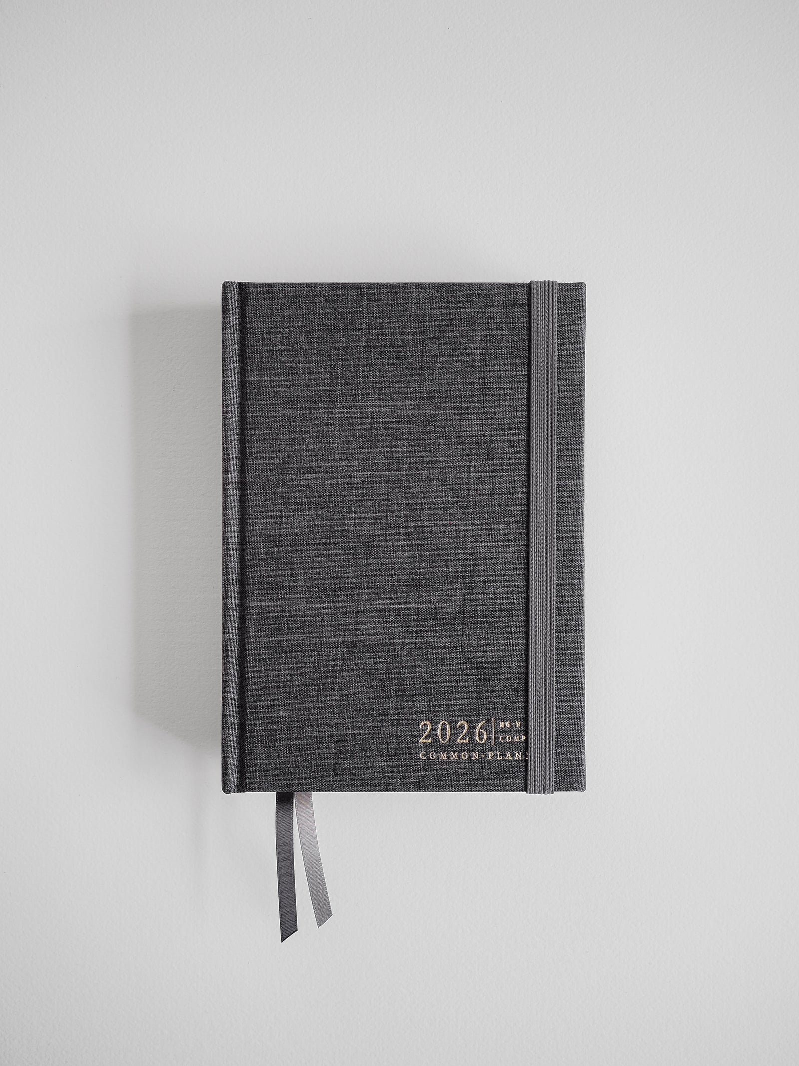 A textured charcoal gray B6 planner for 2026, displaying "Common Plan" branding and finished with a dark attached ribbon.