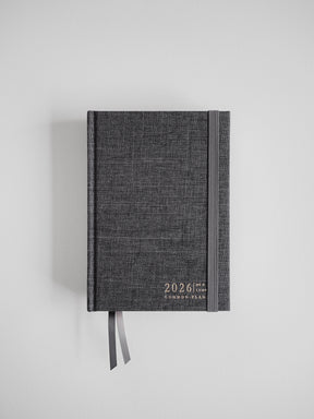 A textured charcoal gray B6 planner for 2026, displaying "Common Plan" branding and finished with a dark attached ribbon.