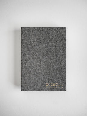 Dark gray, textured B6 daily planner for 2026, featuring elegant gold foil lettering displaying the year and planner title.