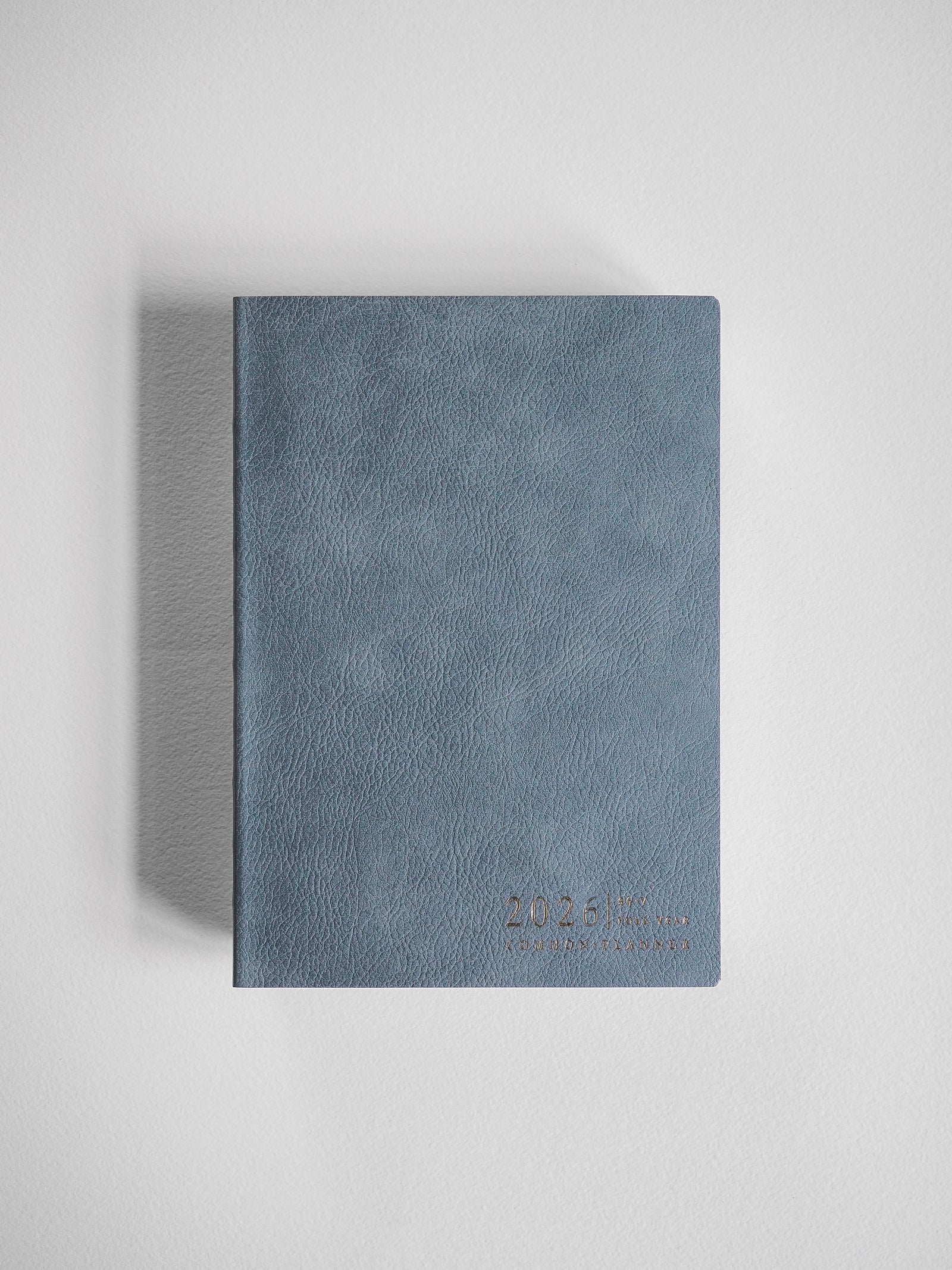 Minimalist B6 vertical full year planner with a textured, muted dusty blue cover, featuring discreet 2026 branding.