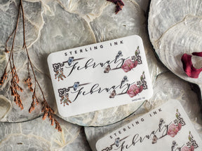 Ornate white sticker reading 'February' featuring romantic, hand-painted floral accents in blush pink, deep red, and muted blue ink.