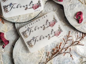 Decorative month sticker for February, featuring the word in elegant script and adorned with delicate, hand-drawn watercolor clusters of pink, blue, and burgundy florals.