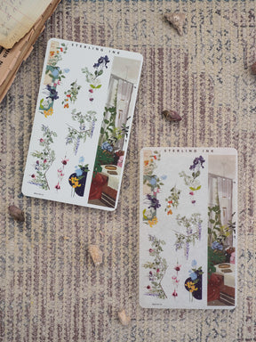 Delicate botanical design featuring clusters of deep purple, yellow, and pink wildflowers set against a charming indoor windowsill scene.