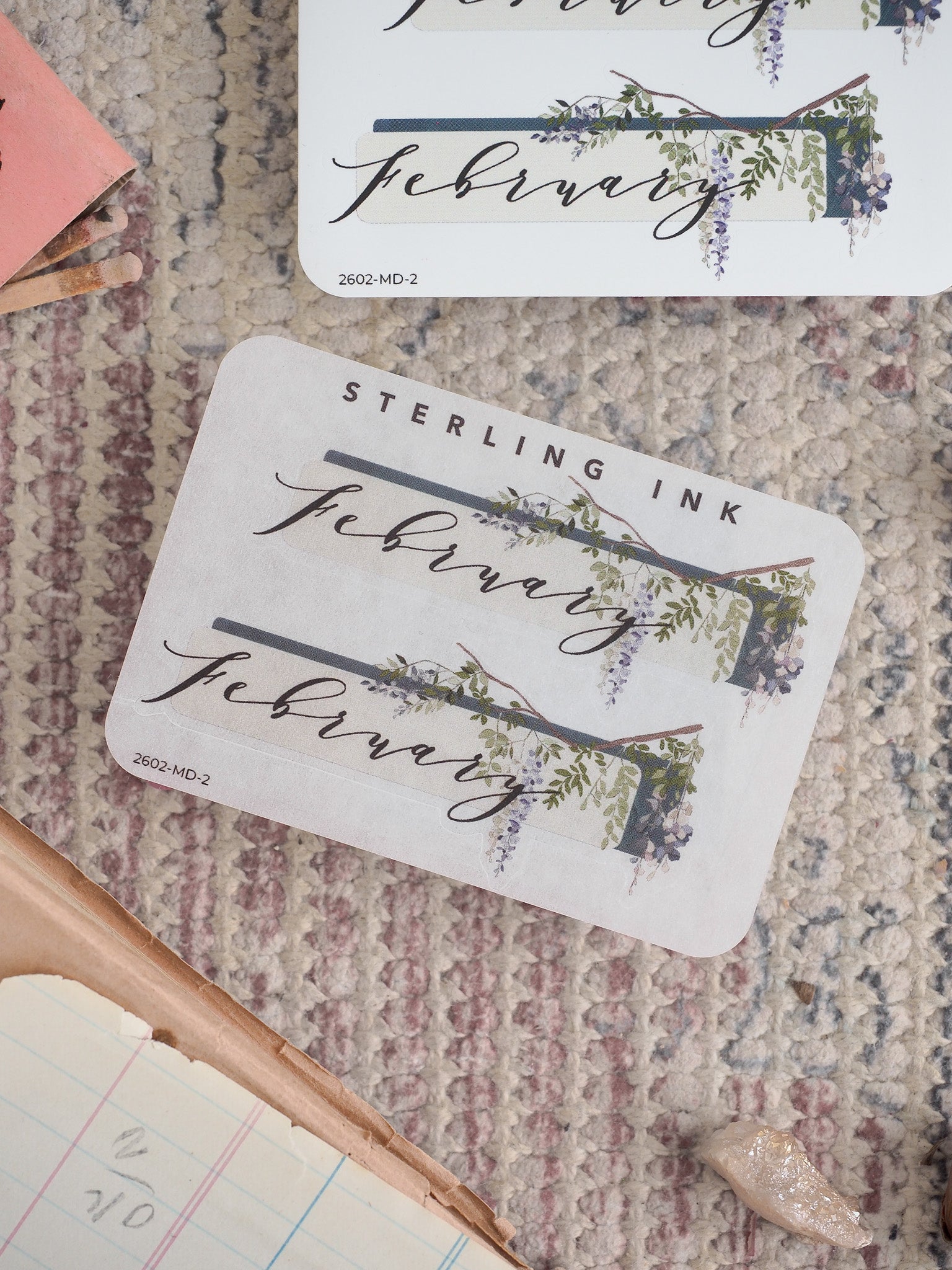 Elegant white sticker featuring "February" typography framed by delicate purple and green floral accents, suitable for journaling and planning.